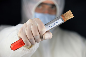 The doctor in a protective suit and mask holding a glass tube with a word COVID-19, during a...