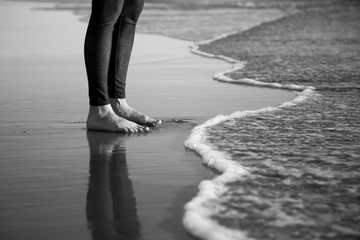 Grayscale closeup shot of human's barefoot legs standing on a sandy beach