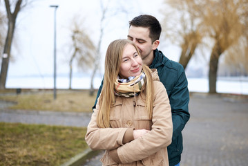 Young brunette man, wearing green jacket, standing behind young blond woman, wearing beige jacket, hugging her, smiling, kissing. Romantic Valentines day celebration