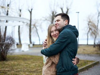 Young couple in love, standing, embracing hugging each other, wearing casual clothes and jeans, on the rainy spring day in city park. Romantic Valentines day celebration outside in town.
