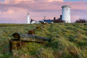 Fototapeta na wymiar Two lighthouses, set in a rural location, with wooden posts leading the eye to the buildings. The sky is pink at sunset