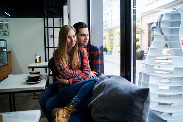 Young couple, wearing red checkered casual shirts, sitting by window in coffee shop. Young blond woman is sipping cappuccino, young brunette man is hugging her. Romantic Valentines day celebration