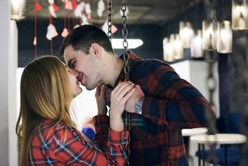 Fototapeta na wymiar Young couple in love, wearing red checkered shirts, sitting on a swing in coffee shop. Attractive brunette man kissing pretty blond woman in coffee shop. Romantic Valentines day celebration