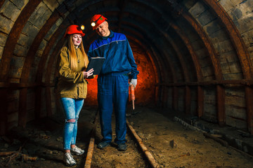 Fototapeta na wymiar A beautiful young girl in a red helmet and with a electronic tablet in her hands is standing with a miner in a coal mine. Business plan discussion. Copy space.