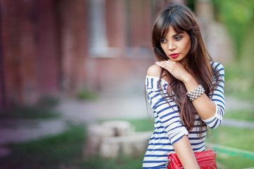 Front view of beautiful lady with long hair holding elegant red purse and looking aside while sitting on bench outdoors. Charming girl in stylish striped shirt with blurred background and copy space.