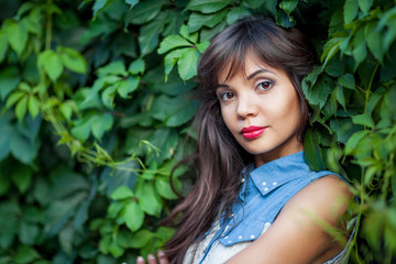 Fototapeta premium Close up of attractive brunette girl posing among green leaves. Charming lady with raspberry lipstick on lips looking at camera and smiling slightly. Concept of female beauty and nature.