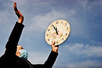 An old grandmother in a medical mask holds a clock against a blue sky. Time is priceless. The old...