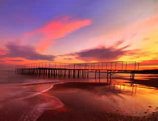 Fantastic sunset on a secluded beach. Old pier and reflection of red clouds in the water.