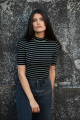 Beauty frontal portrait of a cheerful girl with straight hair, dressed in modern clothes, makeup, supported by a wall.