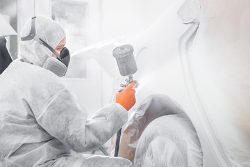 Body painter with airgun spraying white paint on a automobile in car maintenance service paint room.