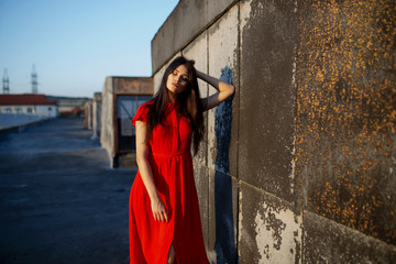 Fototapeta premium Sensual young beautiful woman in red dress, with makeup, have closed eyes dreaming, posing on roof, near old black wall.