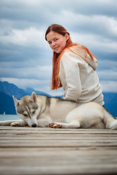 A young woman with brown hair and white sweater is sitting at the pier at the lake with calm water. A Siberian husky female dog is lying down near the girl. The Alps mountains in the background.