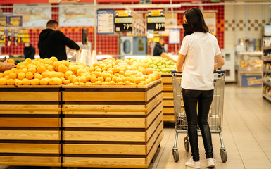 Young woman wearing disposable medical mask shopping in supermarket during coronavirus pneumonia outbreak. Protection and prevent measures while epidemic time. Buys oranges. Back view.