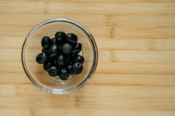 black olives in a glass bowl on a wooden table, top view, closeup