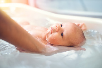 Small cute big-eyed beautiful baby bathes in warm water in the hands of a caring mother. The...
