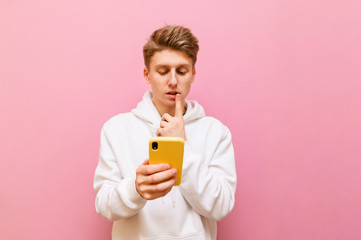 Pensive young man stands on a pink background and uses a smartphone, wearing casual clothes. Teenager in white hoodie uses the internet and thinks isolated on pink. Copy space