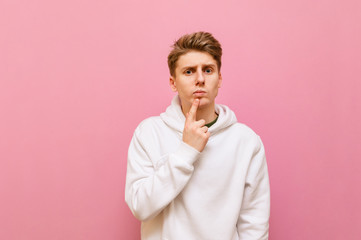 Portrait of pensive guy in casual clothing on a pink background, looks into the camera with a serious face and thinks.Student in a white hoodie put finger to his beard, and thinks.Thoughtful gesture.