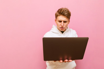 Concentrated young male freelancer working on laptop with serious face, isolated on pink background, wearing white hoodie. Guy uses a laptop on a pink background.