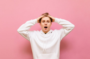 Shocked young man in casual clothes stands on a pink background holding his head, looks in camera...
