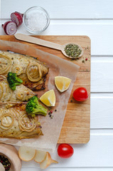 buttery tasty fish mackerel saba with lemon, sliced on filet with tomatoes, onion rings herbs and broccoli in baking paper on a cutting board on a white wooden background