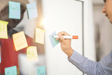 close up of asian businessman employee hand using and placing sticky note on glass board arranging mind map working in modern office, concept of planning, brainstorming business ideas and strategies