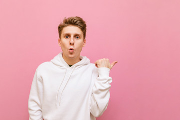 Surprised teenager in white casual clothes stands on a pink background and points his finger away at copy space. Funny guy with a surprised face shows his finger on a blank space and looks in camera