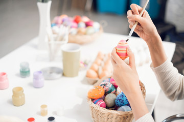 Obraz na płótnie Canvas close up female hand holding easter eggs and paintbrush coloring the eggs in basket working on desk with brushes and paint cartilage, smiling joyfully having fun decoration celebrating easter holiday