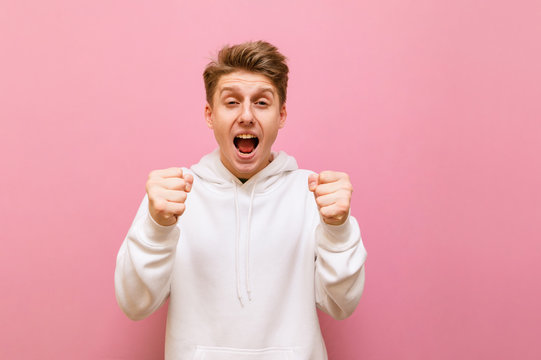 Portrait of joyful guy in white hoodie cheering with fists up on pink background, looking into camera with smile on face. Guy winner isolated on pink. Copy space