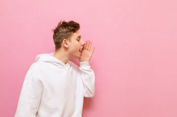 Portrait of a cheerful guy in a white hoodie looks away and shouts with his hand attached. Positive guy isolated on a pink background, put his palm to mouth and speaks