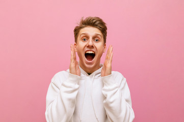 Closeup portrait of shocked guy in white hoodie on pink background looking into camera with mouth open and shouting with joy. Emotional young man yelling WOW, isolated.