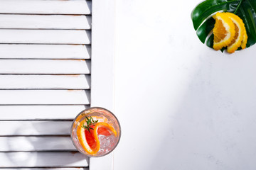 Cold summer drink in a glass with slice of grapefruit and ice cubes on a white background with part of wooden jalousie. Top view.