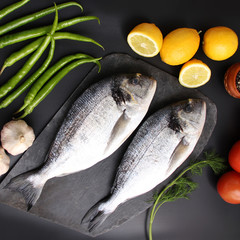 lemons, tomatoes, hot peppers and two carcasses fresh raw atlantic saltwater fish Sparus aurata with shiny silver scales lie on a black background, cooking concept