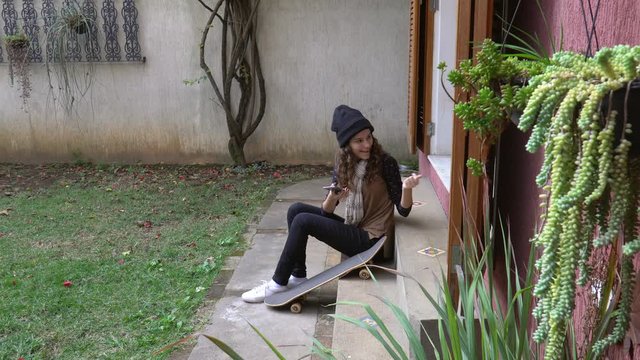 Beautiful fun girl wearing a beanie and scarf sitting on home garden floor with a skateboard calls her black dog to try to take a selfie with cell phone but the dog leaves and she laughs. 4K.