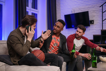 Fierce noisy likable 30s multiracial male friends encouraging with shouts their favourite football team and interfering their male friend to have mobile conversation