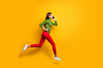 Fototapeta na wymiar Full length profile photo of funny lady jumping high crazy rushing to meet friends speaking over telephone wear casual green jumper red pants isolated yellow color background