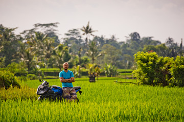 A young man sits on a sports bike and looking into the camera, among the fresh rice field.