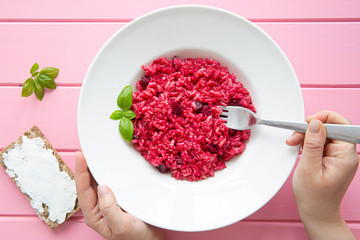 Risotto with beetroot, female hands hold fork, pink wooden background. Top view.