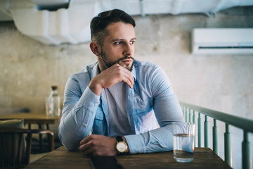 Dreamy caucasian male in trendy shirt and with wristwatch looking away thinking about plans and ideas on break in coffee shop, brunetta 20s hipster guy sitting at table in cafeteria concentrated