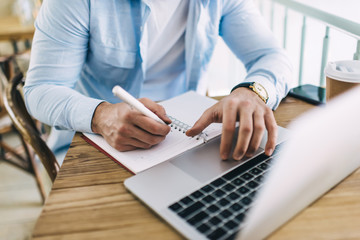 Cropped image of skilled male student writing homework in notepad using laptop computer for...
