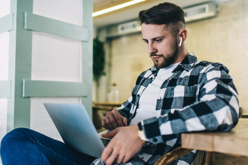 Serious confident businessman in earbuds concentrated on distance job reading mails on laptop computer in cafe, pensive caucasian hipster guy watching video using modern gadgets and 4G connection