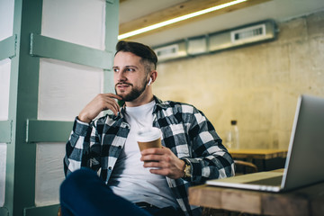 Thoughtful male student in casual wear learning online course using earbuds and laptop computer in...