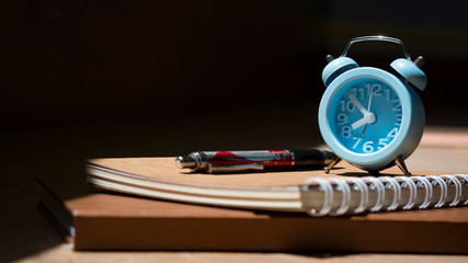 Sunlight and shadow on surface of little blue vintage alarm clock with ballpoint pen and notebooks on wooden table in black background, close up with copy space