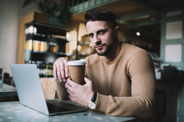 Serious handsome male entrepreneur holding coffee cup looking at camera during remote job process, caucasian 20s businessman enjoying coffee while having freelance project on laptop computer