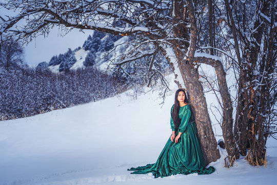 A girl in  fabulous long green dress in the snow-covered mountains. A woman in a fairy-tale dress in the mountains in winter. Winter fairytale photo session of a beautiful girl in Sairam su Kazakhstan