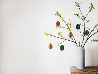 Easter holiday home decoration. Tree branches in vase with fresh spring leaves decorated with festive colorful Easter eggs, white wall background, copy space. Easter holiday preparation, greeting card