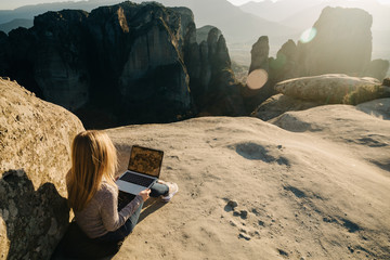 Young blonde woman working on laptop outdoor with beautiful mountain view at sunset. Female...