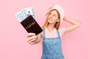 Happy young girl with a hat on her head , with a money fan and a passport isolated on a pink background