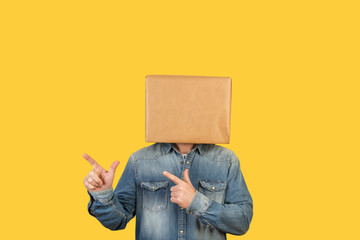 Hombre con caja de cartón en la cabeza con brazos levantados y manos con dedos apuntando hacia la derecha sobre fondo amarillo liso brillante aislado. Vista de frente. Copy space - obrazy, fototapety, plakaty