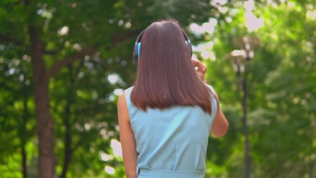 back view woman with headphones on the head listening music walking on the street surrounded green trees