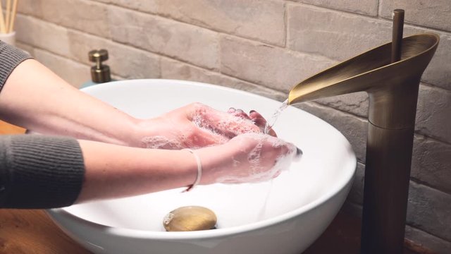 Young woman washing and rinsing hands with soap in a white oval sink bowl and brass fitted tap